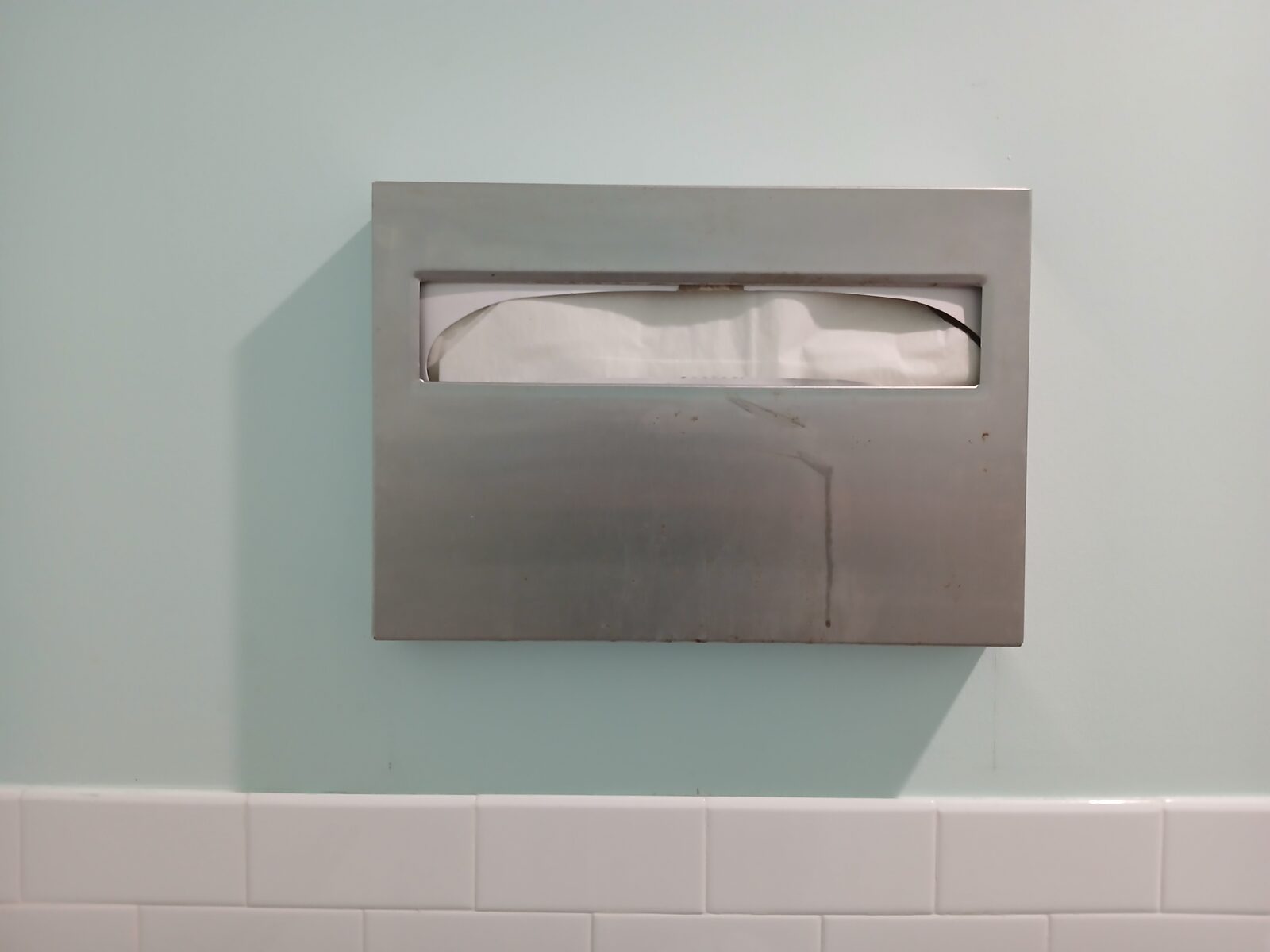 A stainless steel toilet seat cover dispenser mounted on a tiled washroom wall.
