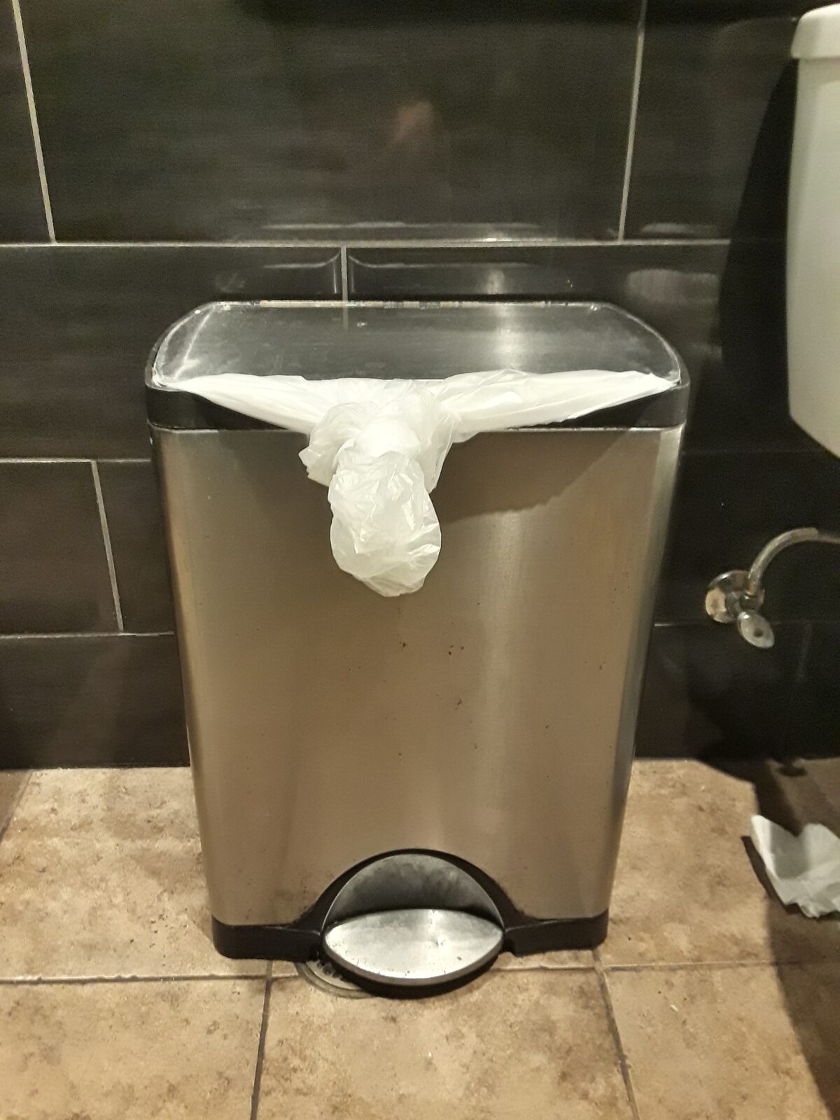 A stainless pedal bin in a washroom with a plastic liner hanging over the rim.